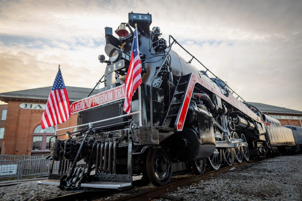 The American Freedom Train No. 1 - a large steam locomotive with two American flags flying in the front - sits outside the North Car shop of the National Museum of Railroad History & Innovation.