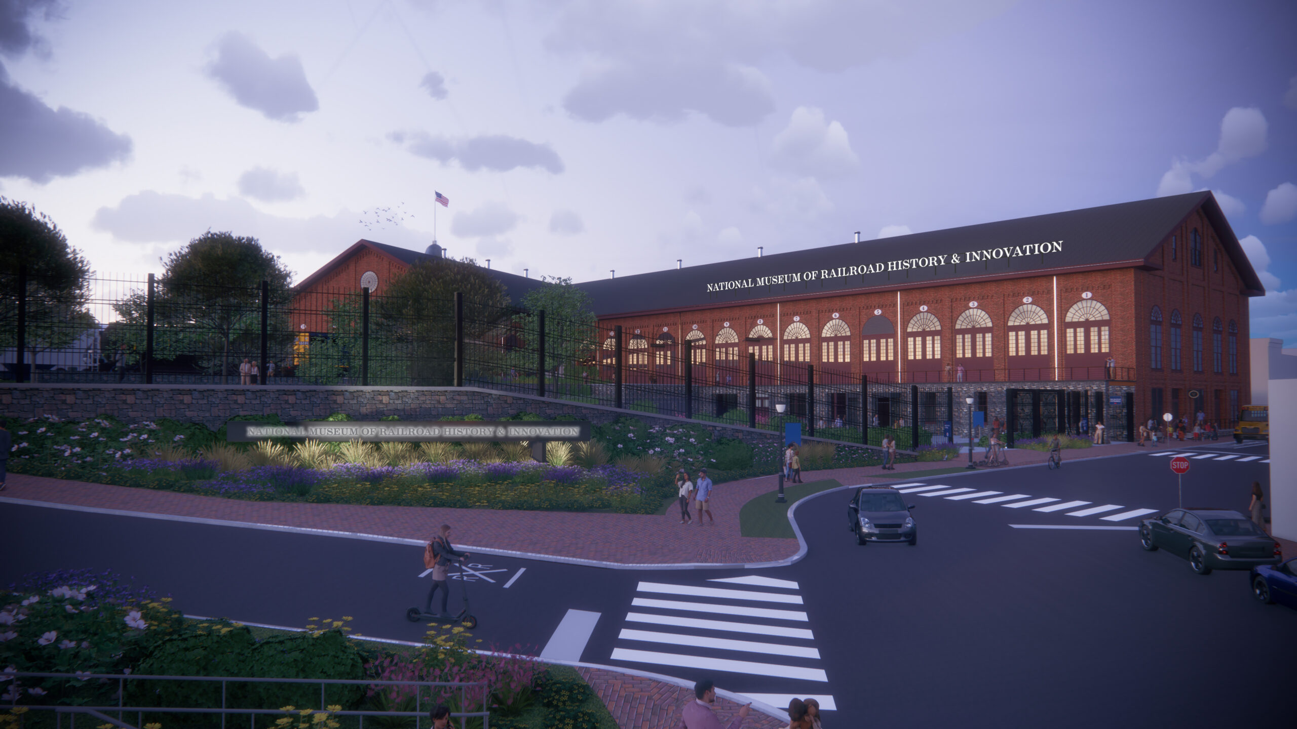 Dusk architectural rendering of the National Museum of Railroad History & Innovation's new entrance, illuminated from within, with the new museum name in lit signage along the roofline and on a low entrance sign, set behind landscaped gardens and a public street.