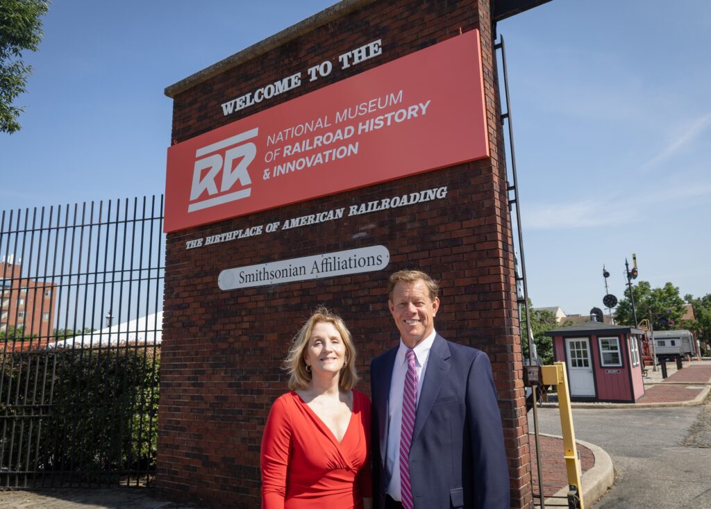 A man and woman stand smiling in front of a brick pillar with a red sign reading "Welcome to the National Museum of Railroad History & Innovation — The Birthplace of American Railroading," with a Smithsonian Affiliations plaque below. The woman wears a red wrap dress; the man wears a navy suit with a pink striped tie. A security shed and railroad signal equipment are visible in the background under a clear blue sky.