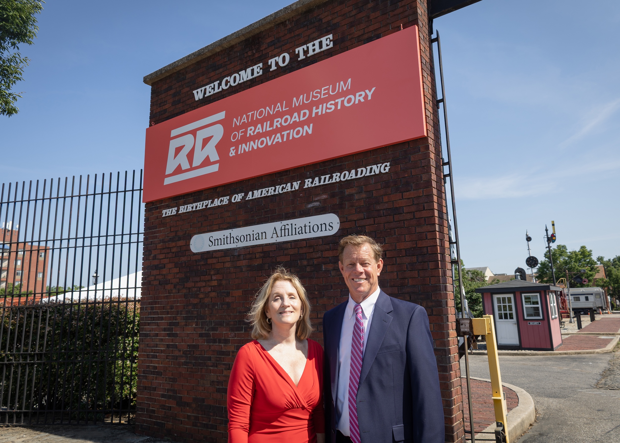 A man and woman stand smiling in front of a brick pillar with a red sign reading "Welcome to the National Museum of Railroad History & Innovation — The Birthplace of American Railroading," with a Smithsonian Affiliations plaque below. The woman wears a red wrap dress; the man wears a navy suit with a pink striped tie. A security shed and railroad signal equipment are visible in the background under a clear blue sky.